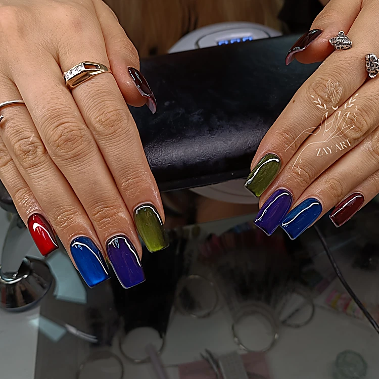 Dos manos sobre el banco negro de la manicura con uñas cuadradas de efecto espejo en rojo, azul cobalto, morado y verde oliva